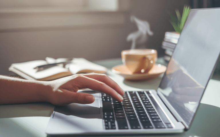 Main d'un rédacteur de contenus tapant sur un clavier portable, avec café fumant et carnet ouvert à côté, illustrant une atmosphère de travail créative et moderne en 2025.