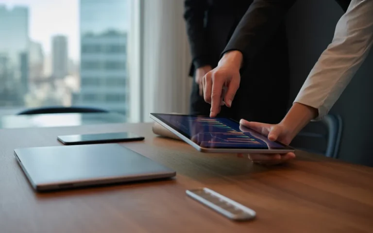 Pourquoi déléguer sa visibilité digitale est rentable: gros plan photo-réaliste de deux paires de mains en tenue business collaborant sur une tablette aux graphiques abstraits colorés, bureau en noyer, open space flou.